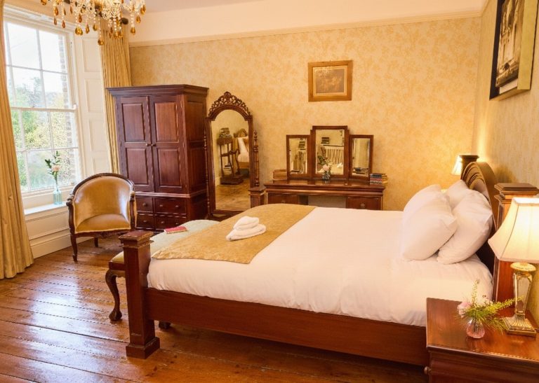 A warmly lit bedroom in Preston House with a wooden floor, a large bed with white linen, a gold throw and towels, antique wooden furniture, a mirror, a lamp, a window with curtains, and a chair by the window.