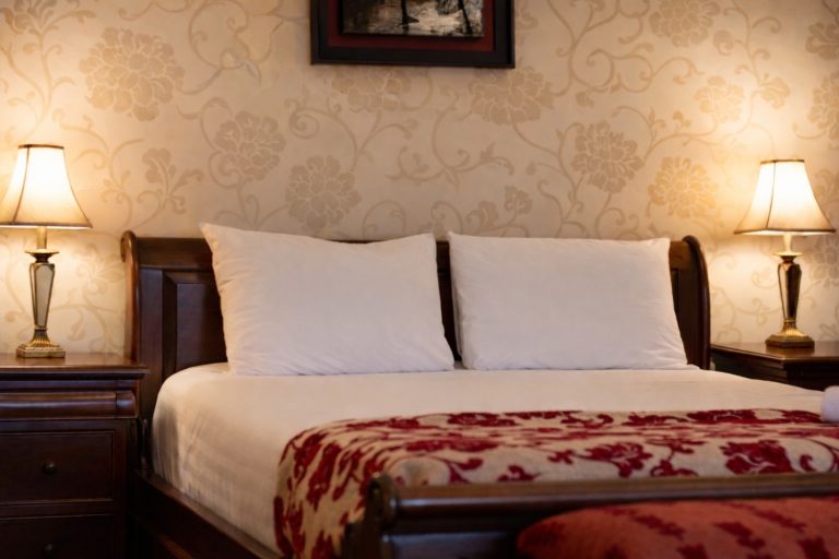 A neatly made bed with white pillows and a red patterned blanket takes centre stage at Preston House, flanked by wooden bedside tables with lit lamps. Beige floral wallpaper and a framed picture complete the inviting scene.