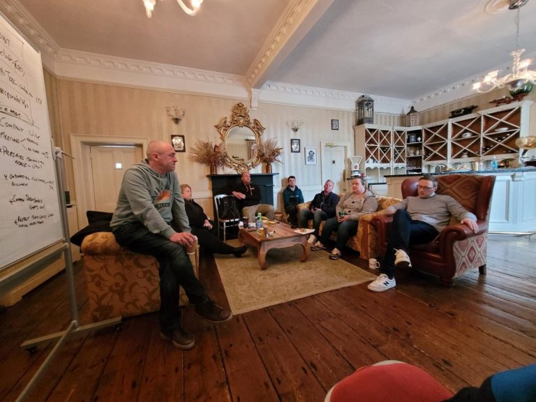 A group of people sit in a cosy, elegant living room at Preston House, with wooden floors and ornate decor, listening to a man speaking while seated on the armrest of a chair. A whiteboard with notes is visible on the left.