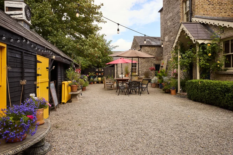A charming courtyard at Preston House with gravel ground, outdoor tables and chairs under umbrellas, fairy lights overhead, potted flowers, and buildings with brick and wooden walls. The setting is cosy and inviting.
