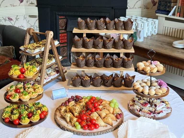 A table at Preston House is set with assorted pastries, muffins, mini quiches, and fresh fruit, displayed on wooden trays and stands, with serviettes neatly placed in the foreground.