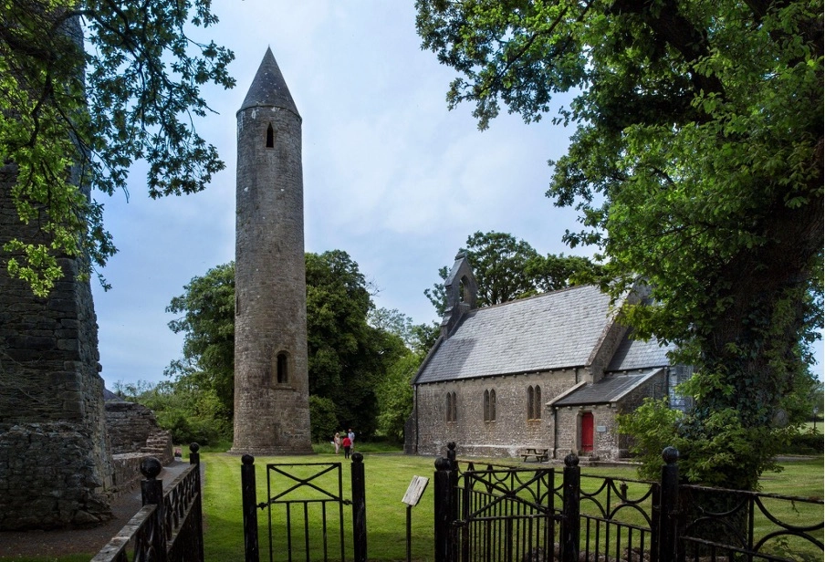 A stone round tower stands beside an old church with a grey slate roof and red doors, near Preston House, surrounded by trees and a black iron fence on a grassy lawn.