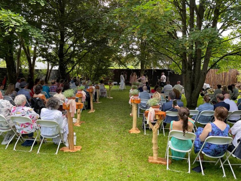 A wedding ceremony at Preston House takes place outdoors under trees, with guests seated on white chairs facing the couple and celebrant. Orange fabric decorates wooden posts along the aisle on the green lawn.