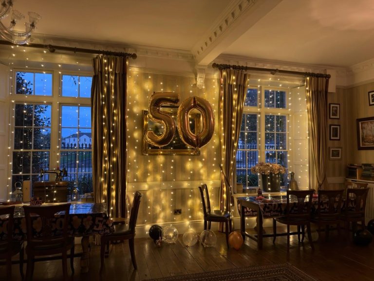 A warmly lit room at Preston House decorated for a 50th celebration, featuring gold 50 balloons, fairy lights, and floor balloons. Tables are set with flowers and party decorations. Large windows reveal a dark evening outside.