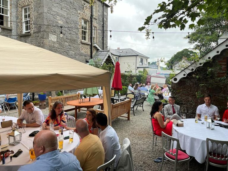 People sit at round tables under a canopy in the Preston House courtyard, socialising and enjoying drinks. The outdoor setting features stone buildings, greenery, and fairy lights overhead on a cloudy day.