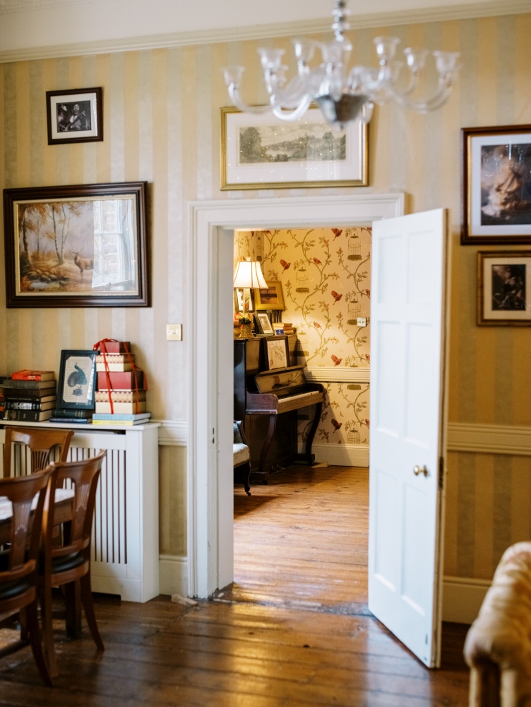 A warmly lit, traditional room in Preston House features wooden floors, striped walls, framed art, and a door open to a cosy space with floral wallpaper, a piano, lamp, and books stacked near the doorway.