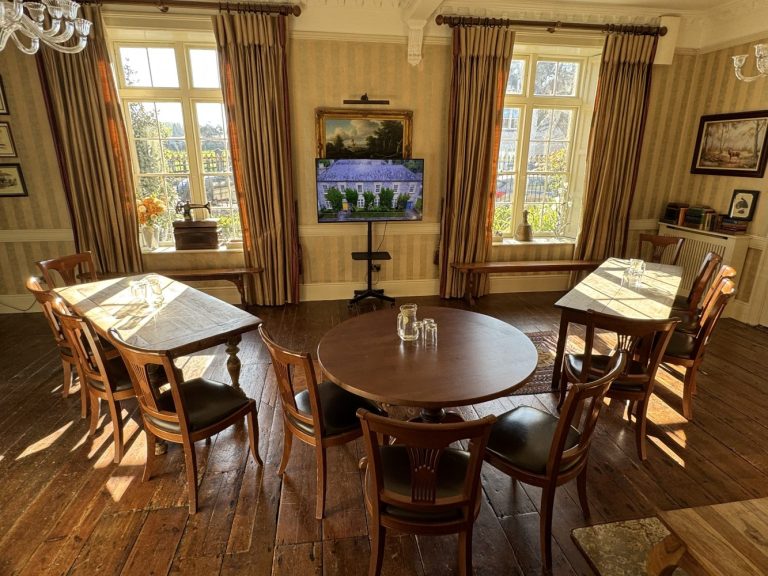 A sunlit meeting room in Preston House with wooden floors, three tables and chairs, a large TV screen displaying a building, tall windows with brown curtains, and decorative items on the walls and windowsills.