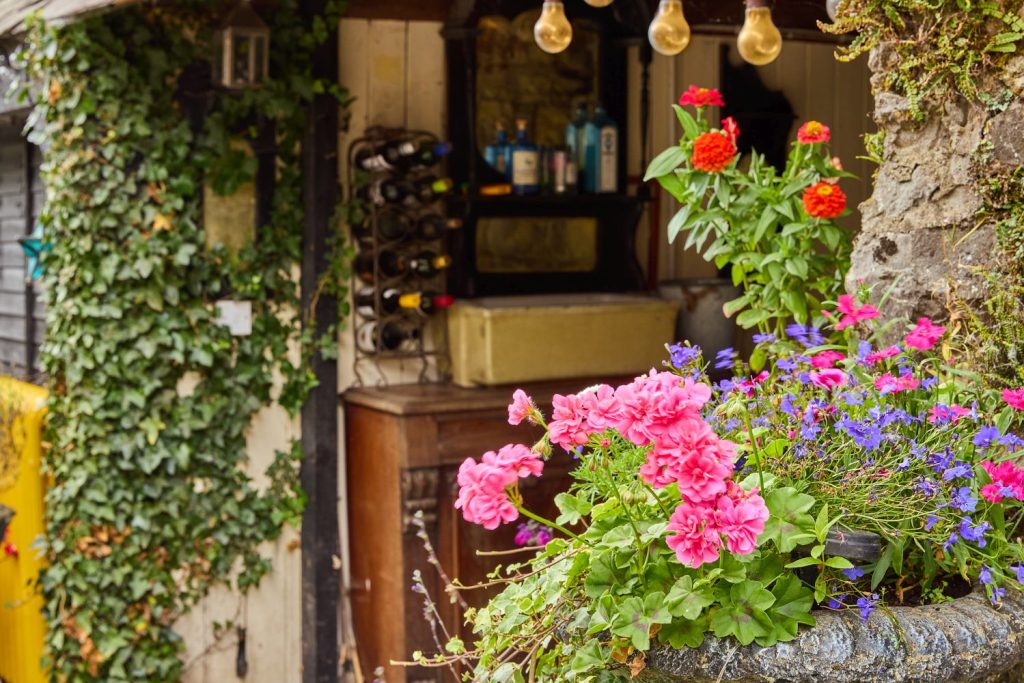 A vibrant planter overflowing with pink, red, and purple flowers sits in front of Preston House’s rustic outdoor bar covered in ivy, with wine bottles and shelves visible in the background.