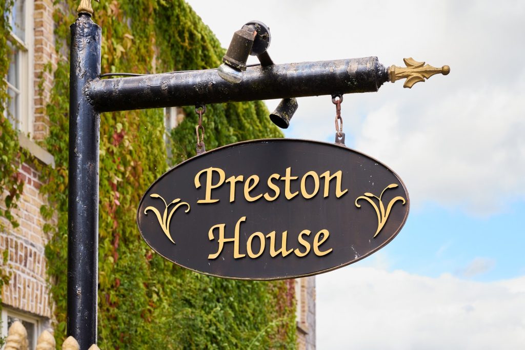 A black oval sign with gold trim and lettering reads “Preston House,” hanging from a decorative metal bracket. Green ivy climbs the brick façade of Preston House in the background under a partly cloudy sky.