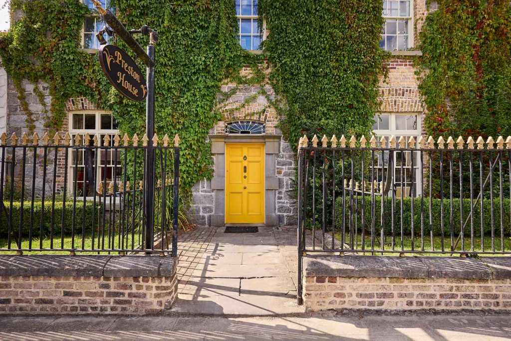 A bright yellow door with a fanlight window is set in a brick and stone house covered in green ivy. Wrought iron gates with gold finials frame the entrance. A black sign reads Preston House.