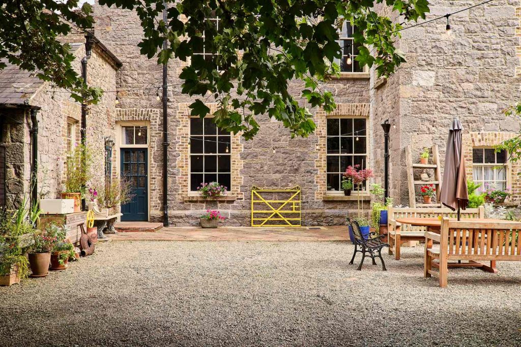 A stone courtyard at Preston House features potted plants, a blue door, large windows, a wooden dining set with a parasol, a small bench, and fairy lights hanging from trees. A yellow gate leans against the building.
