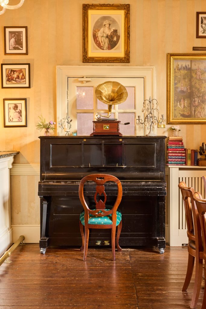 A vintage room at Preston House features a black piano against the wall, an ornate wooden chair in front, a gramophone on top of the piano, framed artwork and photographs on the walls, and books stacked to the side.