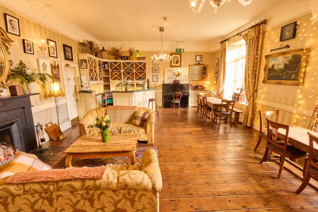 A cosy, sunlit café inside Preston House with wooden floors, vintage furniture, sofas, tables, wall art, and fairy lights. A small bar sits at the back, and large windows fill the inviting space with natural light.