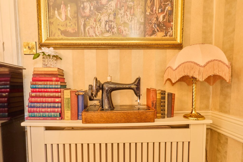 A vintage Singer sewing machine sits on a radiator cover in Preston House, surrounded by stacks of colourful books, a gold lamp with a pink fringed shade, and a framed painting on a striped wallpapered wall.