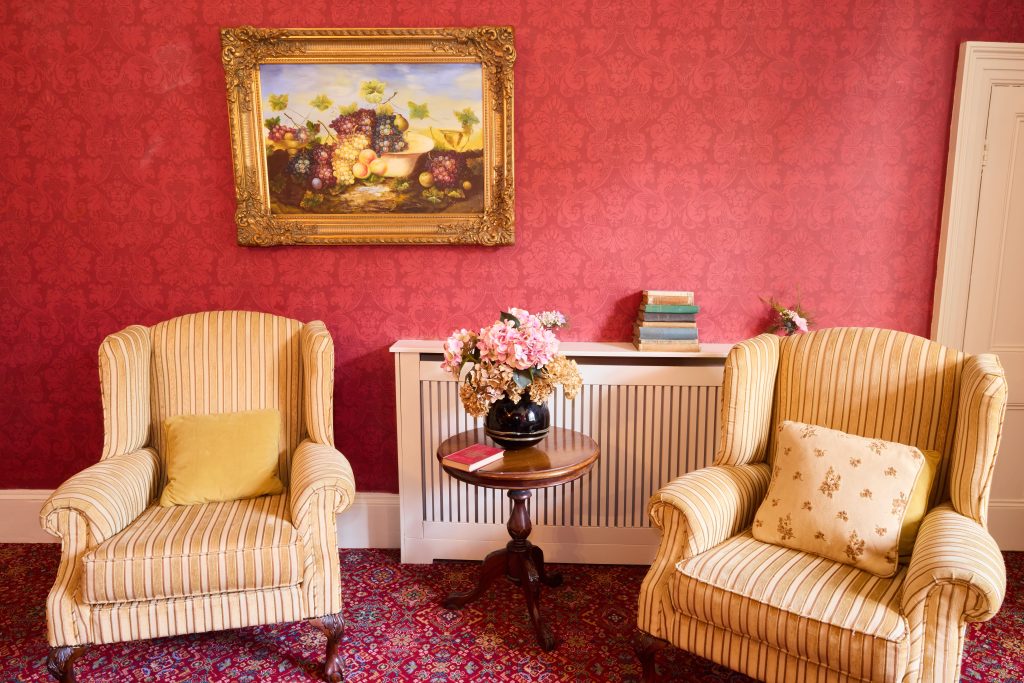 Two striped armchairs with cushions face a small round table holding a vase of pink flowers and a closed book at Preston House, set against a red patterned wall with a fruit still life painting and books stacked on the radiator cover.