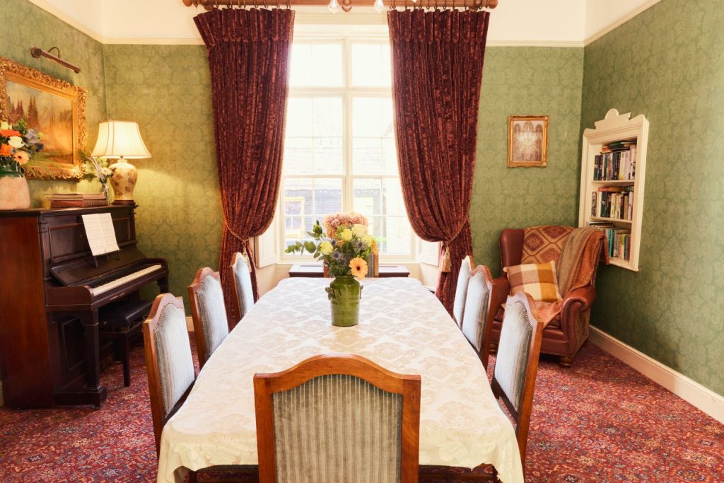 A cosy Preston House dining room with a piano, a table covered with a white cloth and flowers, vintage chairs, an armchair with a cushion, bookshelves, green wallpaper, and large windows with red curtains letting in sunlight.