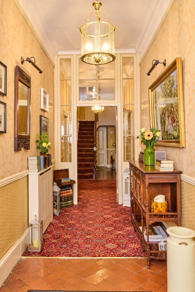 A well-lit hallway in Preston House features patterned wallpaper, red carpet, wooden furniture, framed art, flowers in vases, and stacked books. At the far end, a staircase with a red carpet runner adds elegance to the scene.
