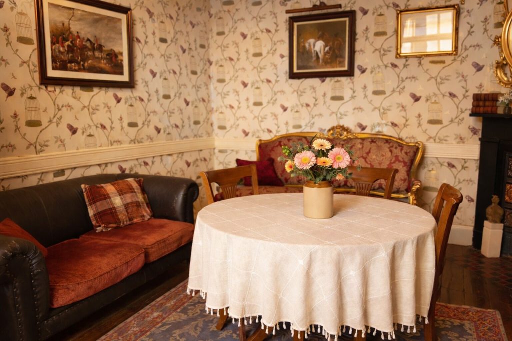 A cosy vintage living room in Preston House features floral wallpaper, a round table with a flower vase, a red upholstered settee, a dark leather sofa, and framed paintings. Warm lighting creates a welcoming atmosphere.